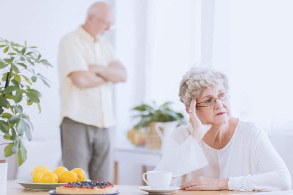 Elder woman sitting at the table, angry because of the argument with her husband standing behind her