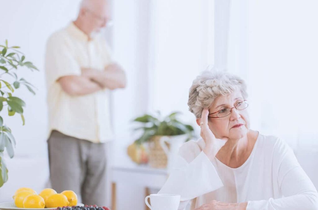 Elder woman sitting at the table, angry because of the argument with her husband standing behind her